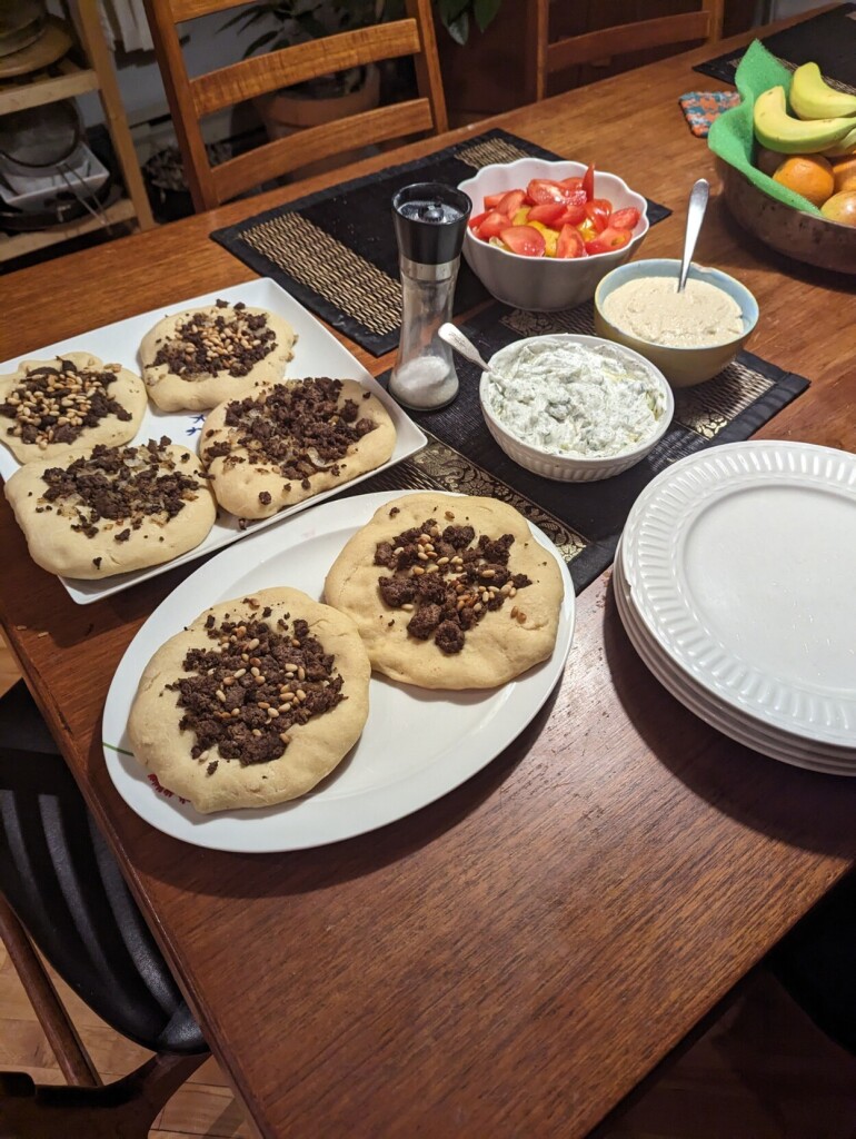 Six Arabic meat pies on two platters, one square and one elliptical. To the side, a bowl of tzatziki, a bowl of hummus, and a bowl of salad with tomatoes and lettuce and peppers. To the other side, a stack of plates.