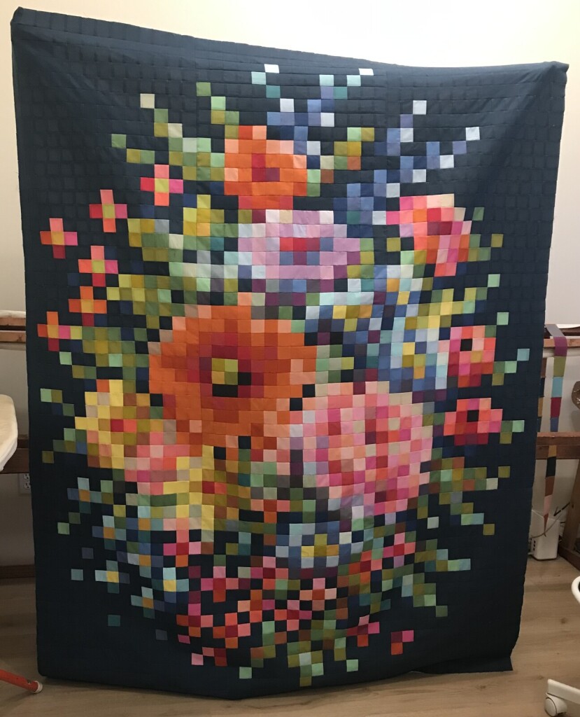 A photo of a pieced quilt top being held up by a person entirely obscured behind it. The quilt top is made entirely of 2”squares pieced together and looks like a cross stitch grid pattern of flowers in a bouquet with greenery behind all on a navy background. The squares are impossibly tiny and represent an intense amount of sewing and work done carefully over a long period of time. 