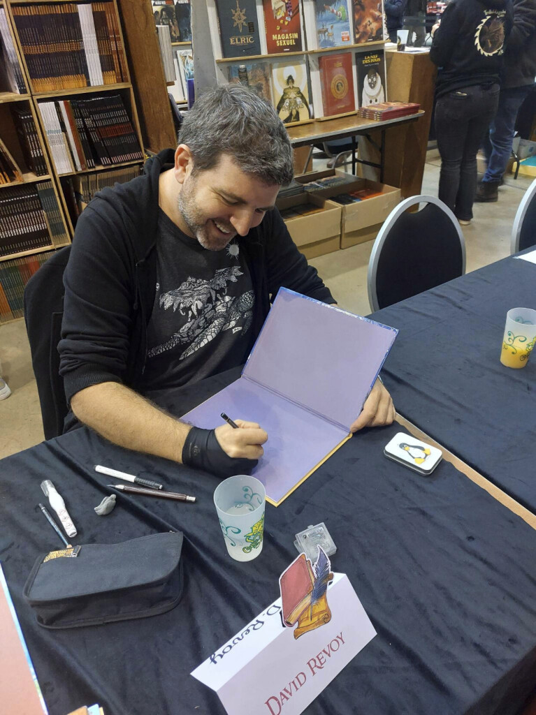 A photo of David Revoy, made from the point of view of a visitor at the start of having the book four of Pepper&Carrot signed at Trolls & Legendes Festival, in Mons Belgium, 2023. 

It's a top-down photograph in color made from a smartphone of a seated white person around 40 years old smiling and happy, with a short beard and a mix of brown and white hair. He wears a tee shirt with the great Great A'Tuin (Discworld, T.Pratchett), and also wears a glove for drawing (synthetic, black) and starts to doodle into a book open. The book is Pepper&Carrot four by Glénat, with its dark violet colors in the introduction. The table has drawing material on it (Tipp-Ex, Pierre Noire, white markers, and a kneaded eraser), a little box with a tux sticker on it, a glass of water, and the table are covered with fabric in black. A label "David Revoy" is on the front to indicate the booth to the visitor. In the background: the library shop of the Festival with many comic books.

Photo CC-By 4.0 Steph Vesch.