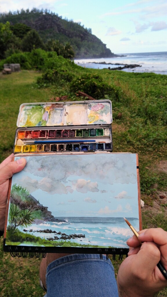 A photo of the coast of Saint-Joseph at Réunion island, under, my hand holding a sketchbook, watercolors, and a painting of the coast.