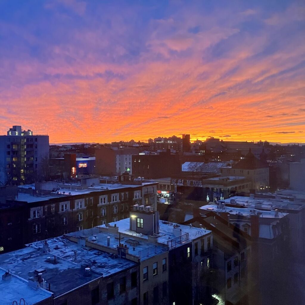 The Brooklyn skyline shot from the ninth floor at dusk. The sky is a radiant graduent of dark orange to vivid purple while the building below have a dark blue shade.