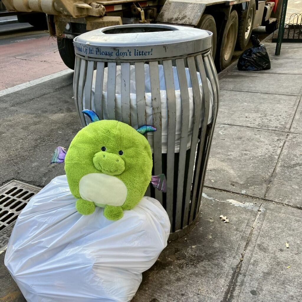 A green round dragon plushie sits atop a white garbage bag against a public street trash bin.