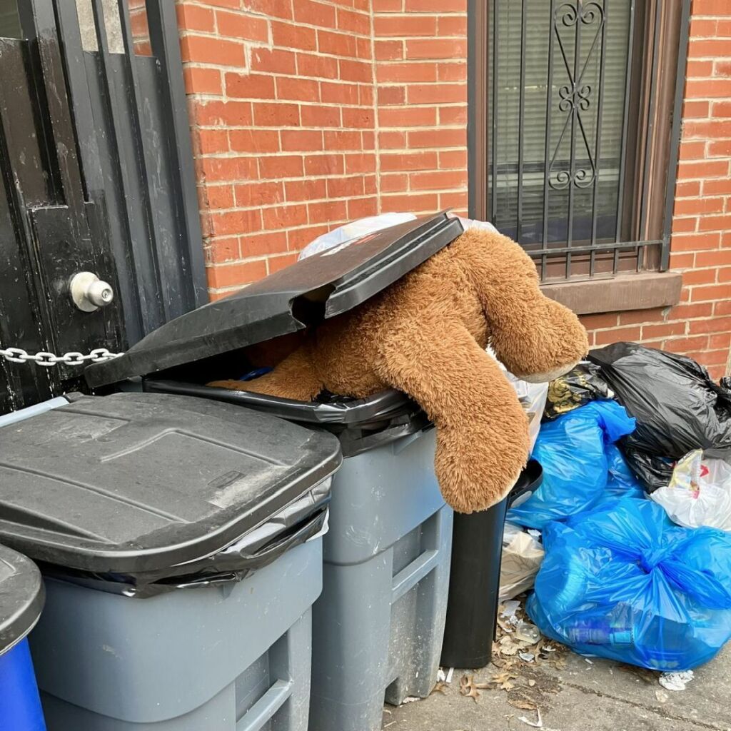 A large brown Teddy Bear is tucked halfway inside a rolling trash bin on a sidewalk as if it was rummaging through its contents.