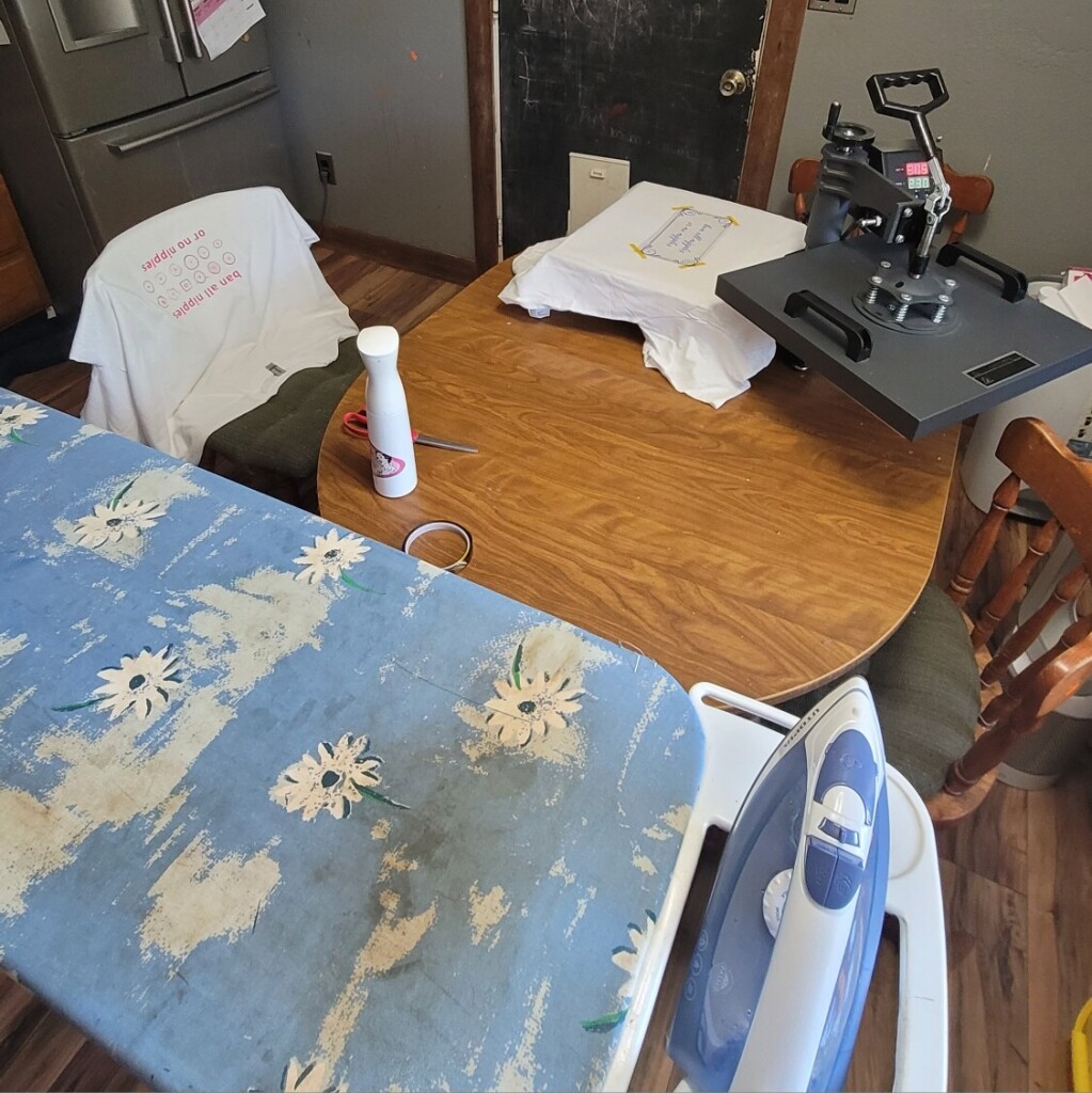 photo of my heat press setup on the kitchen table, with an iron and ironing board, shirts cooling, and the heat press itself