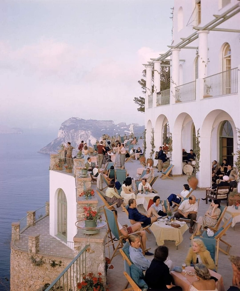 A terrace at a cafe in Capri, Italy, 1949.
Photo credit: Ralph Crane.

A group of casually dressed people lounging on an elegant terrace overlooking the sea. The building is white stucco with arched windows and white pillars. 