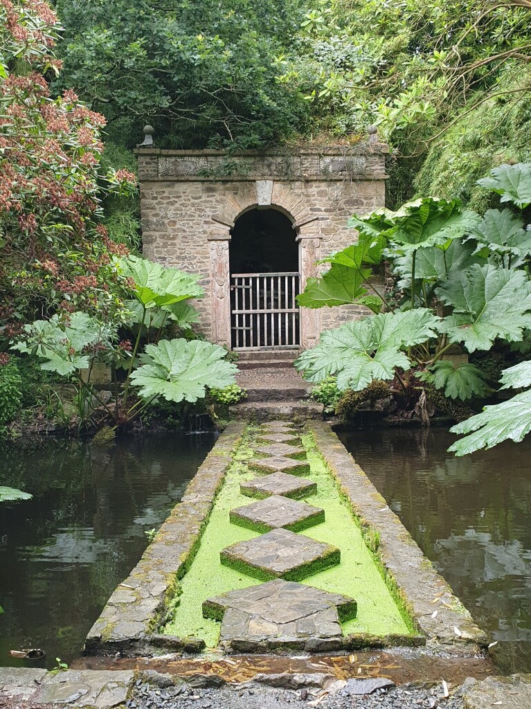 a way of stones and duckweed leading to a weathered stone gateway. around the way is a pond. also giant plant leafs are around the gate.