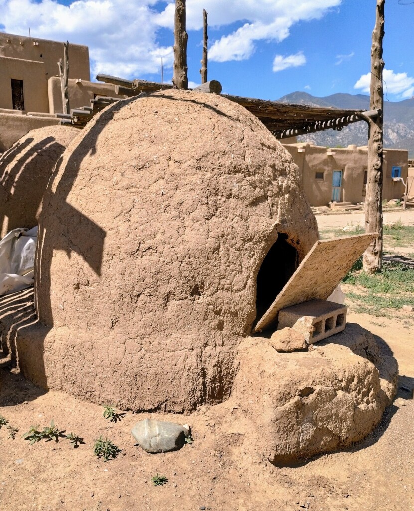 A horno is a large domed adobe outdoor oven. This one has a small wood panel that is slightly open over the opening to the oven. The horno is a ruddy clay color. A large Pueblo is in the distance behind the horno and everything sits below a blue sky with a few scattered white clouds.