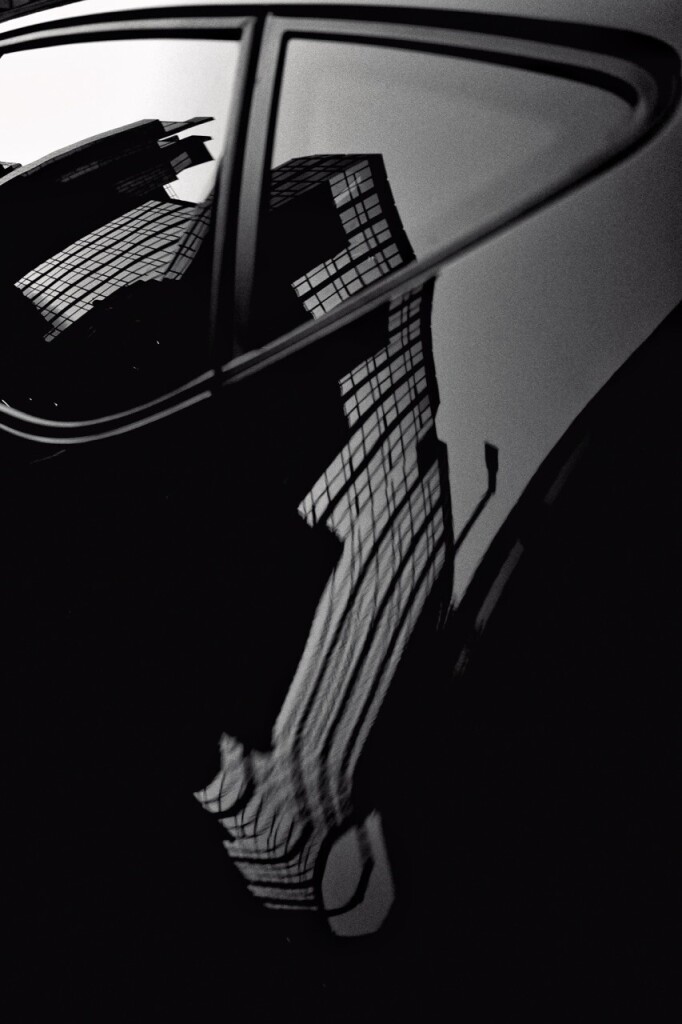 Black and white portrait format image showing the warped reflection of a city office block in the window and bodywork of a black car (mine!)