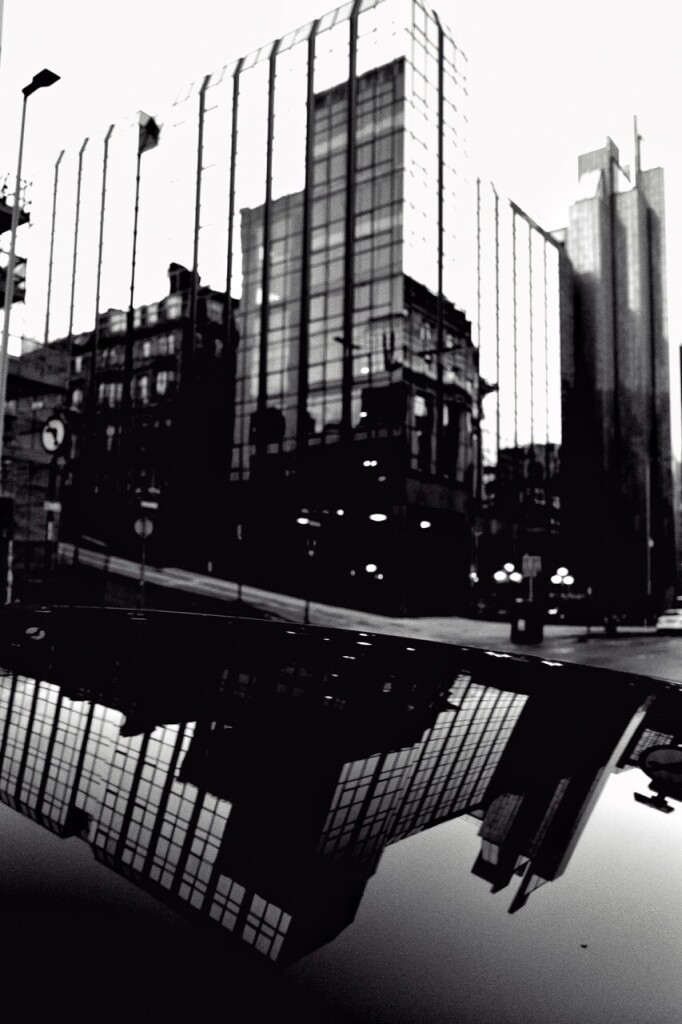 Black and white high contrast portrait format image showing a glass fronted office building in which can be seen the reflections of other office buildings across the street, and the whole scene is reflected in the shiny black roof of a vehicle parked on the street