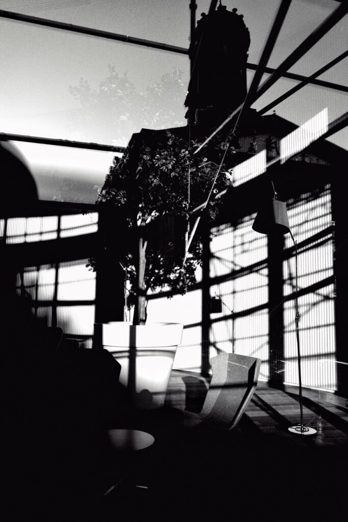 Portrait format, multi-layered black and white image shot into the window of an office complex where there is a tall potted tree, a lamp, a table and chair. The shadows of scaffolding which surround the building can be seen in the far wall, while the reflection of the Glasgow Gallery of Modern Art and some of the scaffolding can be seen at the top of the image.