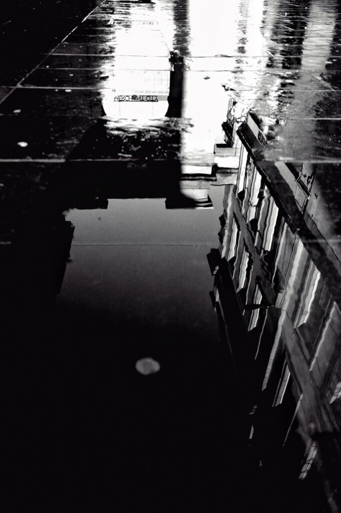 Black and white portrait format image taken after heavy rain, showing the reflections of buildings and the archway leading to Exchange Square in a large, long puddle