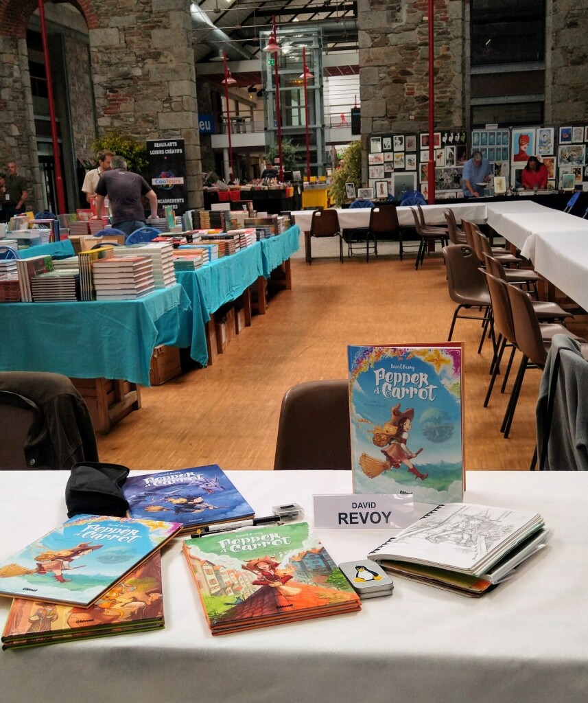 A photo of the main hall in "Bulle a croquer " festival in Saint-Brieuc, France. It's morning and I'm one of the first to get tools ready on a table in foreground. I have my pens, the four Pepper & Carrot books and a sketchbook. In background, the bookstore still prepares all the books. Time to take a coffee ☕ :-)