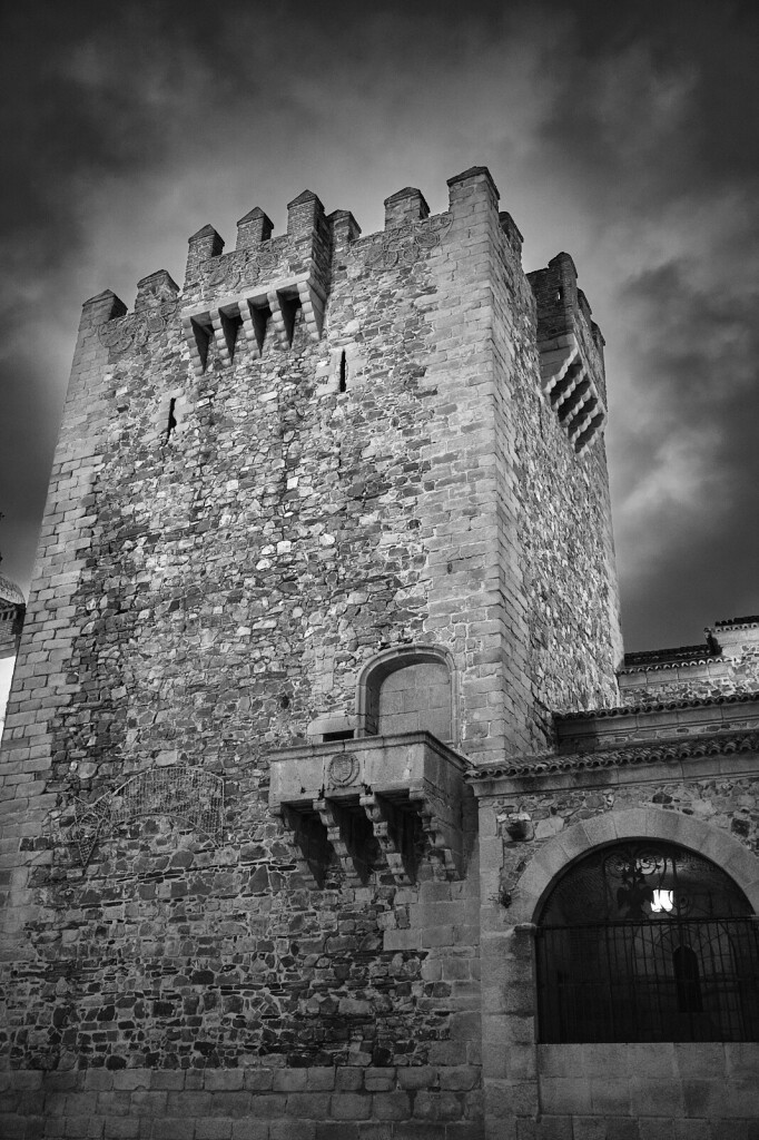 An evening photo of a medieval fortification tower. This sits on the plaza mayor in Caceres, Spain. It is lit with artificial light, and the stones that compose it give it great texture and tonal variety. There is a cloud above that produces an odd effect. 