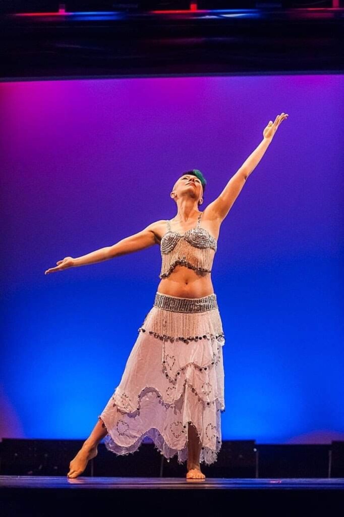 Belly dancer with turquoise hair and a beaded white and silver dance costume stands with arms extended. 