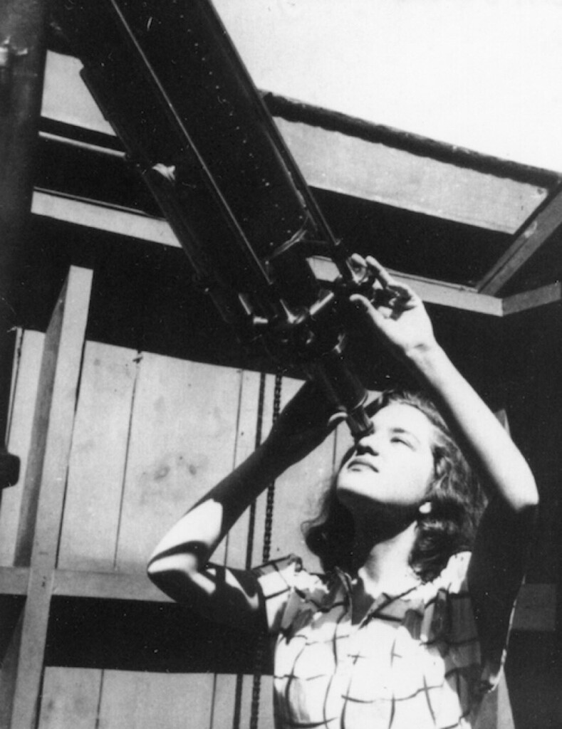 Black and white photo of a young Vera Rubin – likely in her teens or twenties – looking through the eyepiece of a long telescope. She wears a blouse or dress with short sleeves and a grid print. Her wavy hair is shoulder length, and there is a wall behind her composed of two-by-fours and wood boards. Her right hand touches the eyepiece, and her left hand is turning a small adjustment knob on the side of the telescope.