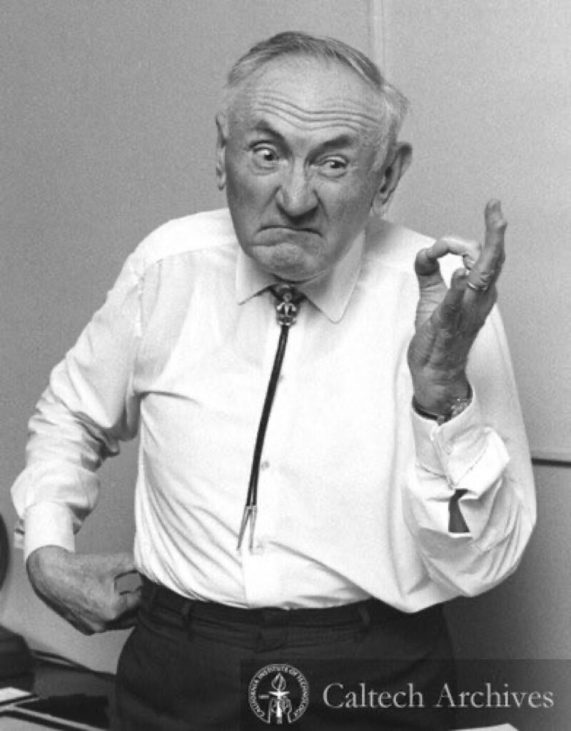 Black and white photo of Fritz Zwicky. He wears dark pants and a white, button down shirt. A tank top undershirt is visible underneath. His right hand is on his hip and his left hand is held up, making an “o” shape with his thumb and index finger. Zwicky, who appears to be in his 60s in this photo, is making a funny, frowning face, like he smelled something bad. He is also wearing a bolo tie, which is a point in his favor.