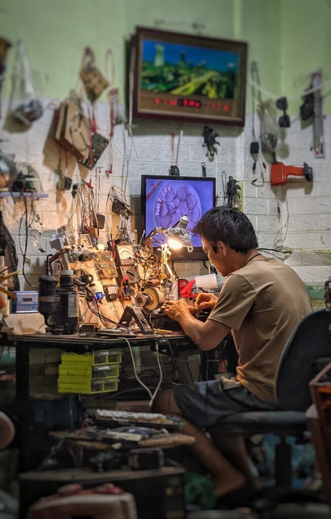 An early evening photo of a young man sitting at a work bench in his electronics repair shop on a back street of Hanoi. He is wearing a brown t-shirt and grey shorts, and we are back and to the left of him so that we appear to be looking just past his left shoulder onto his work. While we cannot clearly see what it is he is working on, we can see the well-used array of meters and tools both on the desk and pinned to a slanting panel at the back of the top surface of the workbench. In the out-of-focus foreground, we see stacks of items. The walls are a seemingly endless series of pegs and hooks upon which hang tools, parts, and unidentifiable bags. Chaotic as the scene is, it is immediately evocative of any number of one-person workstations we will have encountered over the years, i.e. everything is in its place and granularly indexed by the only person for whom it ultimately matters: the present technician.