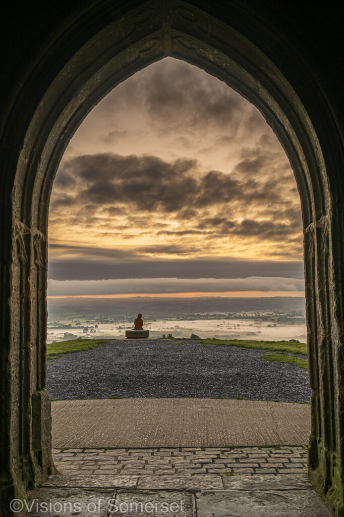 Through the archway is a person watching sunrise. Cloud layers in the sky. Mist below across the land.