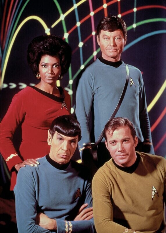 Promo-Bild Star Trek: vorne sitzen Spock mit verschränkten Armen und Kirk, dahinter stehen Uhura mit der Hand auf Spocks Schulter und McCoy. Der dunkle Hintergrund wird mit bunten Lichtbögen belebt.