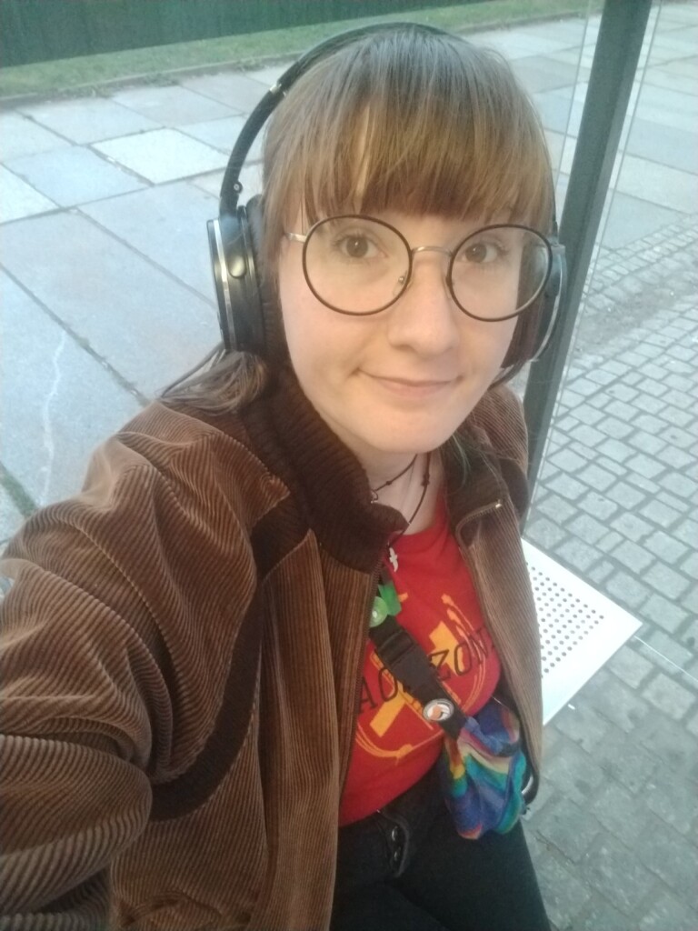 Ich, klein, blass, rotbraune Ponyfrisur, große Brille, braune Jacke und rotes Chaos-Zonen-Shirt, sitze an einer Haltestelle