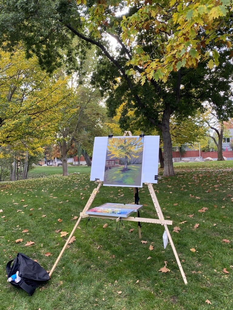 A plein air easel with a pastel painting of a yellowing tree in a park 