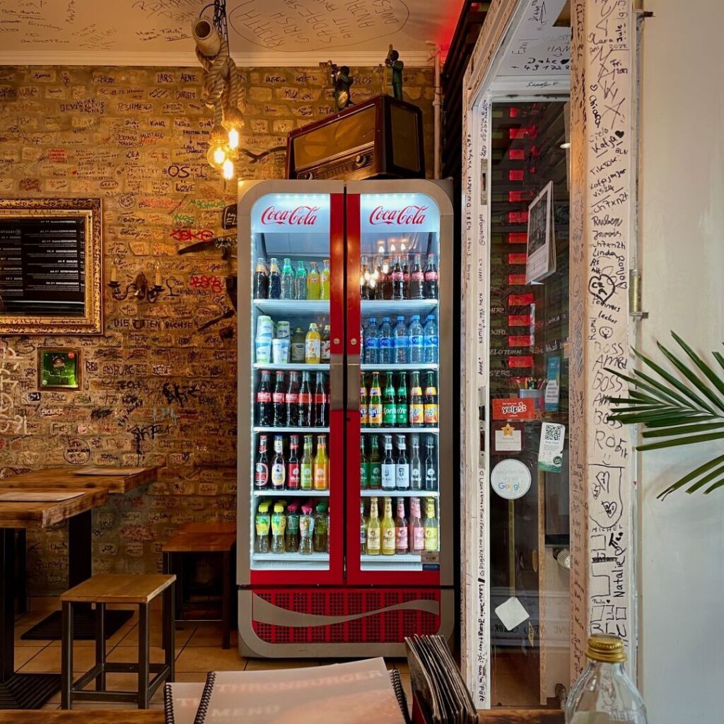 sitting in a burger restaurant, opposite another table, a large refrigerator with an old radio on it, the entrance door to the right