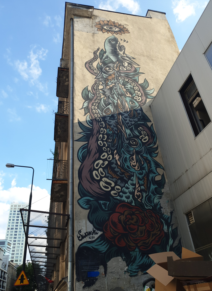 Photo of a high building during construction works (protection nets attached to it front wall) with large mural on side wall. There is dense set of fish and octopus tentacles, with shapes like leaves and fish fins sticking from this mass. There is giant dark red rose flower in bottom part and yellow eye with red petals around above top part.