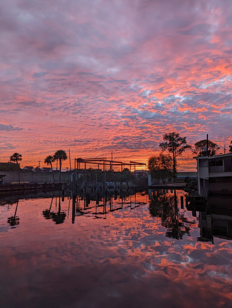 A vibrant orange-pink sunset against a blanket of tufted cloud. The sky is reflected in nearly still water with some trees and urban decay nearly silhouetted across the horizon.