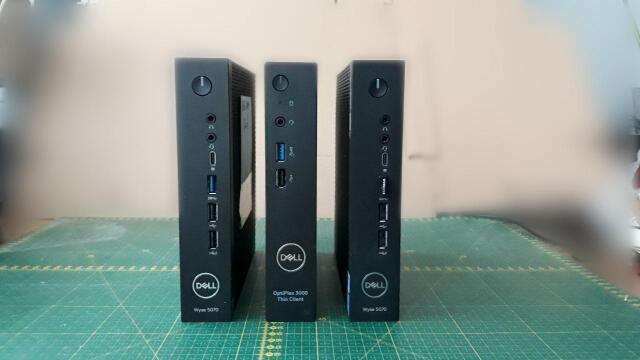 Na stole stoją pionowo obok siebie trzy komputery typu Thin Client: dwa Dell Wyse 5070 a w środku Dell Optiplex 3000