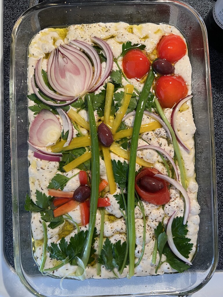 Uncooked focaccia dough in a glass baking dish topped with an artistic arrangement of vegetables including cherry tomatoes, red onion slices, green beans, bell pepper strips, black olives, and herbs, ready for baking.