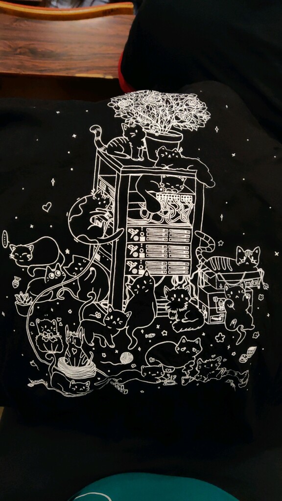 A photo of the t-shirt from La Contre Voie, a white on black whimsical drawing of LOTS of cats playing around in a server rack