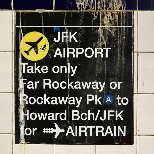 A New York subway pillar sign informing travelers of the correct train to take to the John F. Kennedy airport. Inside the plane logo on a yellow disc, a paper ant was glued to the sign.