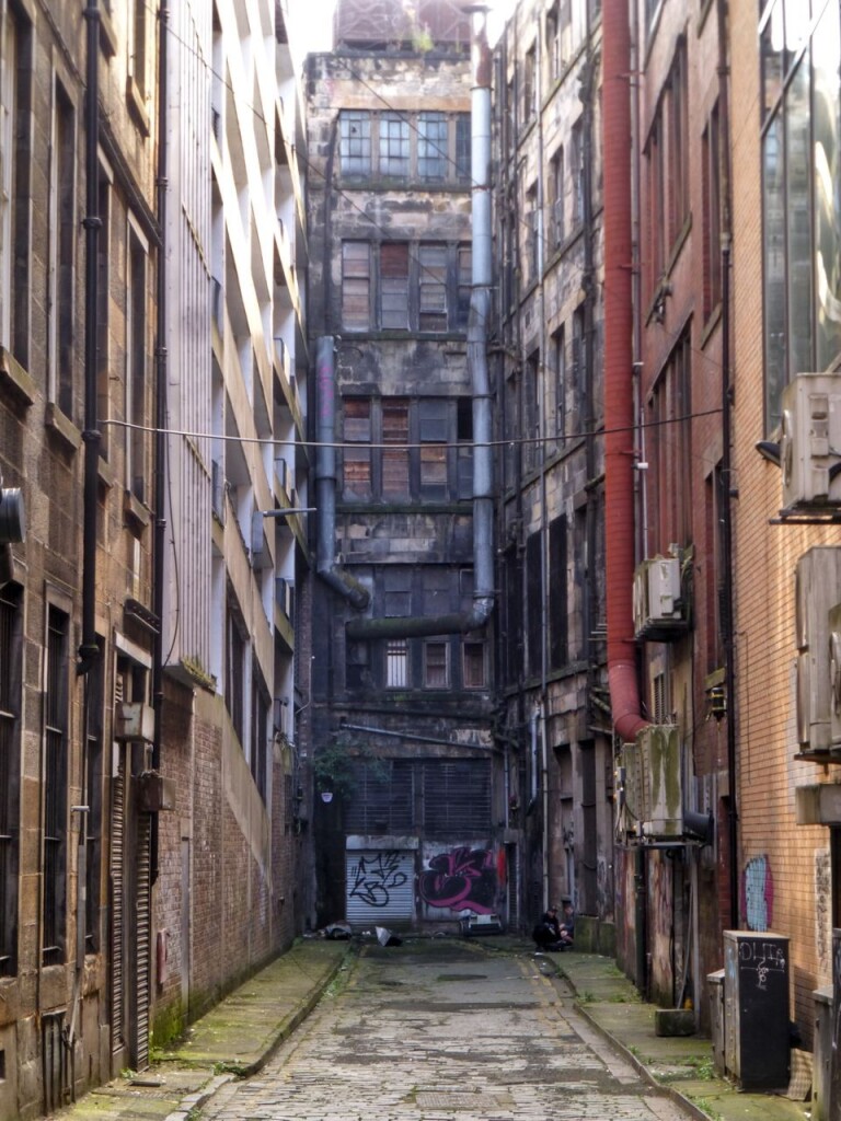 A narrow cul-de-sac, the road surface uneven cobble stones,  enclosed by tall buildings with lots of ventilation ducts, boarded-up windows, windows protected with bars, a few aircon units, and graffiti. 

The focus is a window in the end wall on the second floor, you can somehow see what looks like daylight shining through it from the other side of the building.