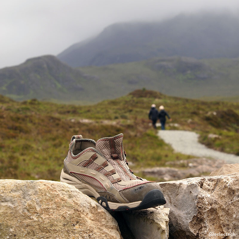 A square photo of some misty Scottish mountains. In the foreground there is a stone wall with a single walking boot, clean but with no lace, wedged between two stones. It is shades of cream and brown, matching the stone of the wall rather well. In the distance, slightly out of focus, there is a path winding through some grass with a couple with matching white hair and dark waterproofs walking away towards a mountain shrouded in a cloud.