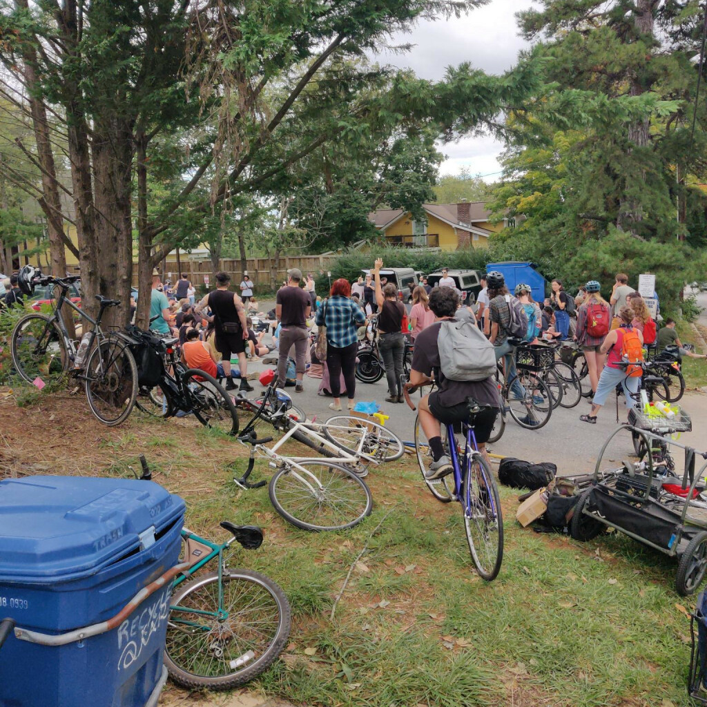 A photo of a large crowd assembled in the parking lot behind Firestorm Books for a community meeting. Participants sit or stand in a circle. In the foreground, several bikes lie in the grass or lean against a tree.