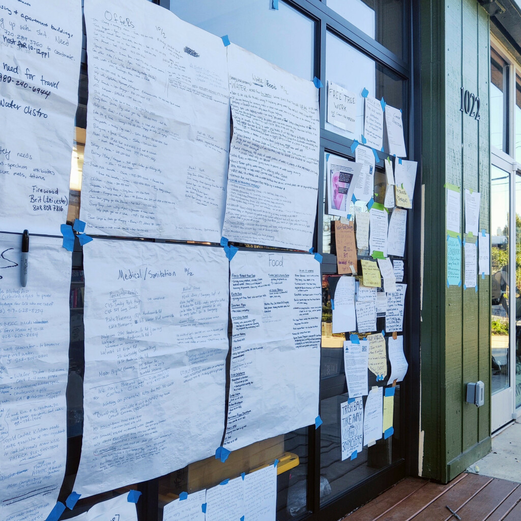 A photo of a roll up door on the front of Firestorm Books covered in sheets of paper with notes about Hurricane Helene and ongoing relief efforts.