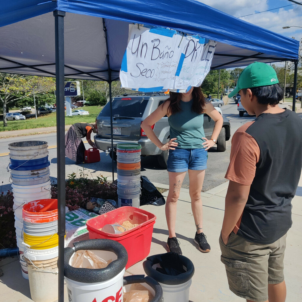 A photo taken outside Firestorm Books shows a pop-up tent full of buckets and sawdust used for making dry toilets. A youth converses with the tent operator, whose face is blocked by a sign in English and Spanish.