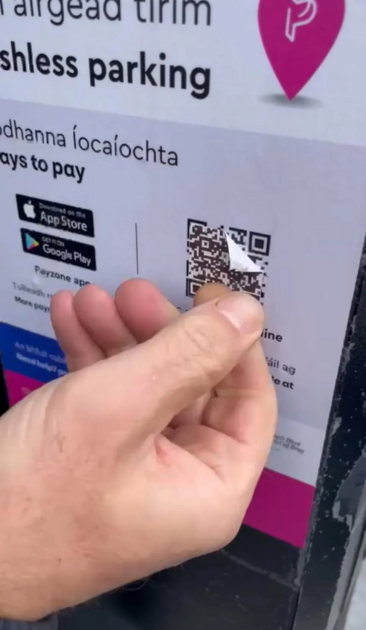 A car parking sign, a QR code has been overlaid on top on the one printed on the sign, a hand is visible in the image, peeling the fake QR code off.