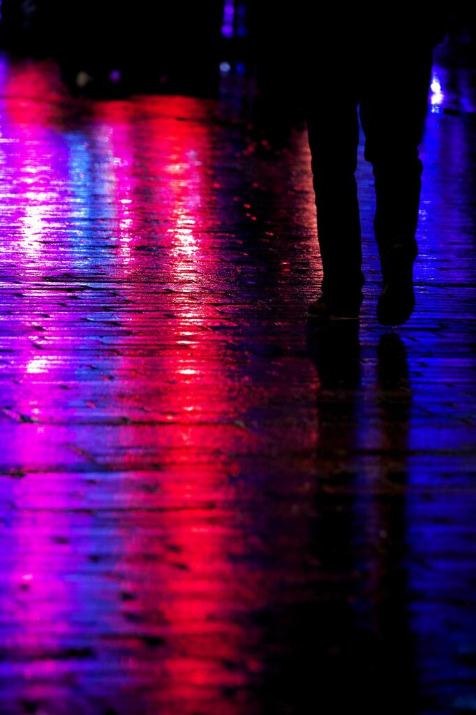 Colour photograph taken in a heavy night-time rainstorm showing the lower half of a lone man walking down a wet street on which are seen the reflections of red, blue and purple neon lights from a carnival.