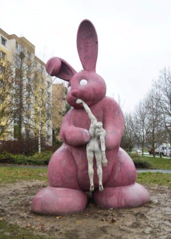 Gray sky, highrise prefabricated housing blocks. On a muddy lawn a giant pink concrete rabbit, holding a white human body. Headless body. Now, the bunny bites a hand. An obvious reference to Goya's “Saturn devouring his son”. Sculpture in Plzen, Czechia, by Adam Trbušek.