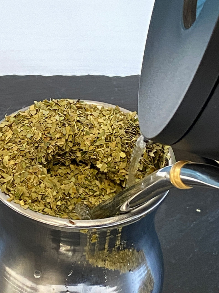 Hot water being poured over green yerba in a silvery recipient (mate).