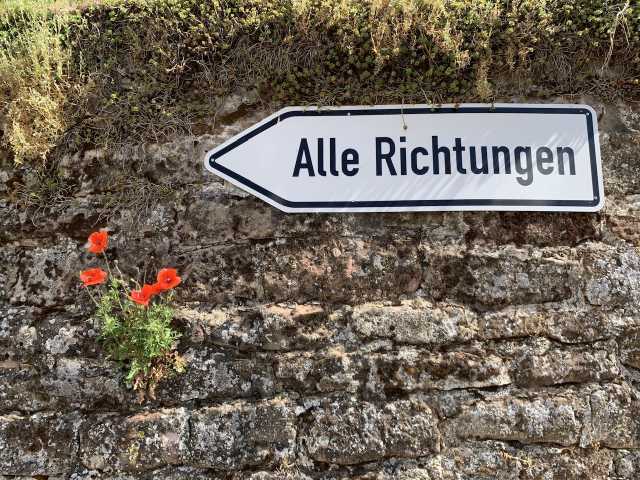 Foto einer gemauerten Wand aus ungleichmäßigen Steinen, die mit Flechten bewachsen sind. Oben an der Mauer, an den Steinen festgeschraubt, ist ein Straßenschild. Auf dem Schild steht „Alle Richtungen“, die zu einem richtungsweisenden Dreieck abgerundete Seite des Schildes zeigt nach links. Links darunter wachsen einige voll erblühte, rote Mohnblumen aus der Mauer.
