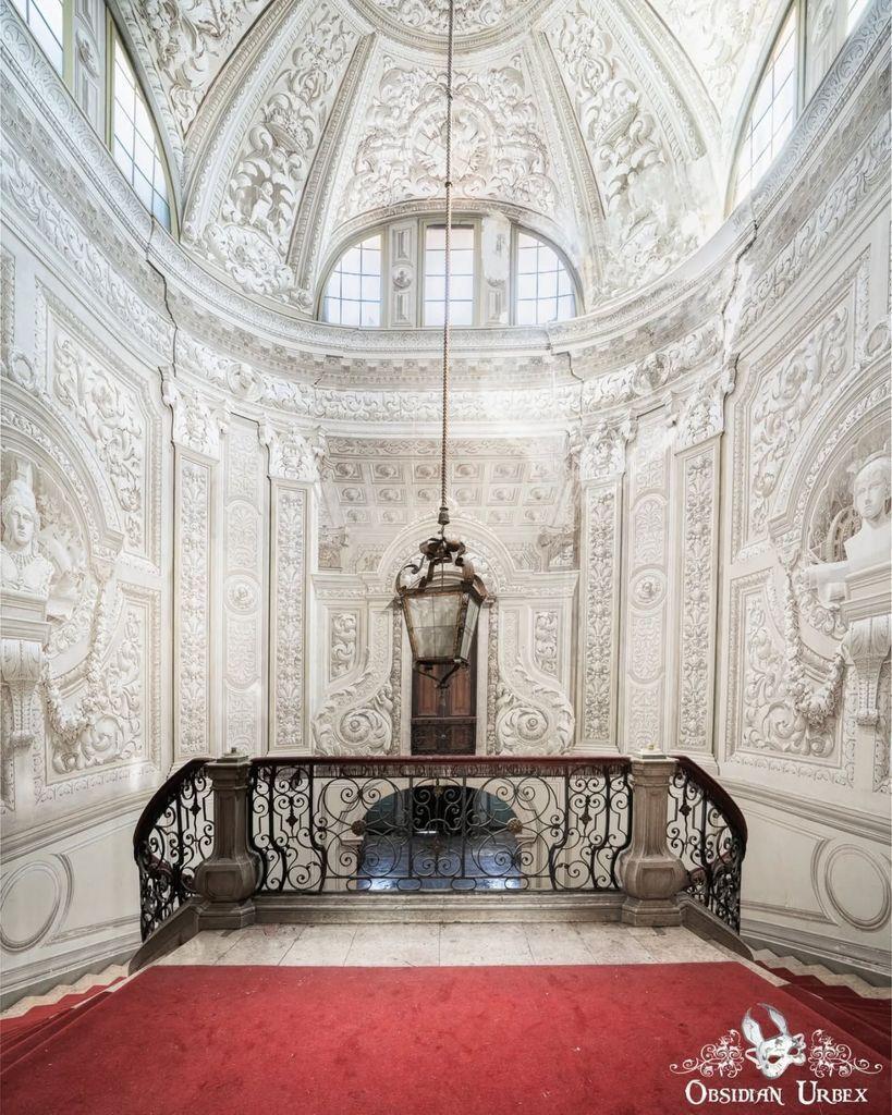 tall room with painted walls to look like 3d scenes. there is a black lantern hanging, the stairs have a red carpet 