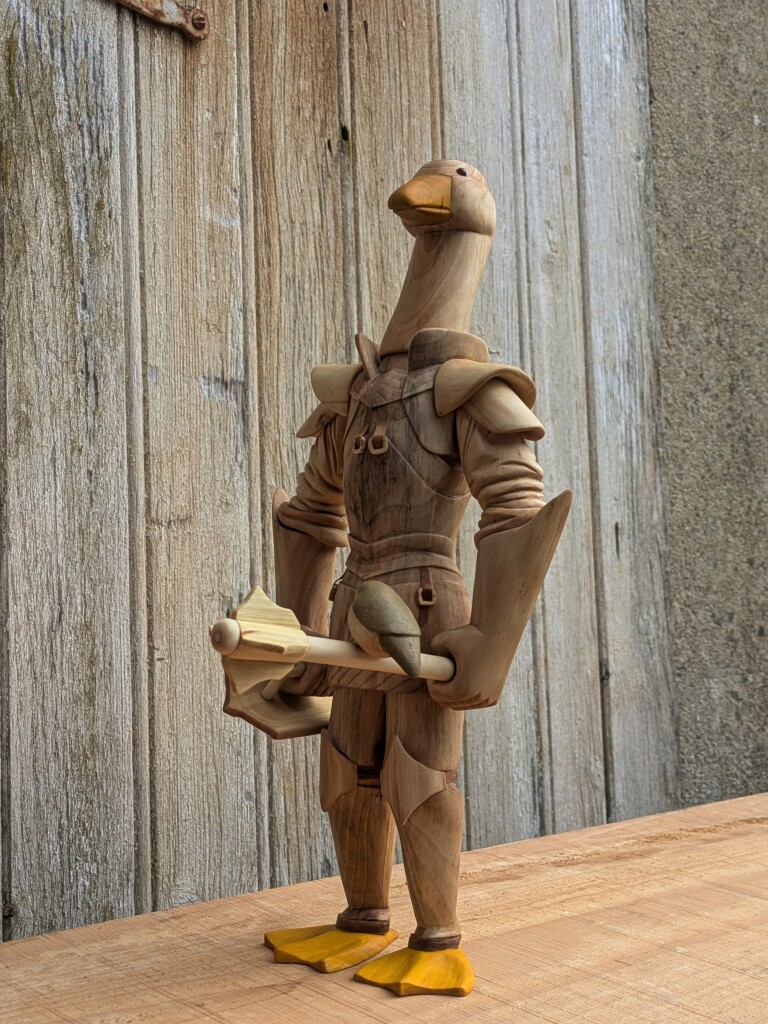Woodcarved humanoid goose wearing plate armour. She's holding a shield and a flanged mace. On the handle of the mace, a red robin landed. The eyes of the goose are made of glass. The feet and the beak are painted in burnt yellow, as the breast of the red robin. The upper part of the robin is painted in brown/grey  and its eyes and beak in black.