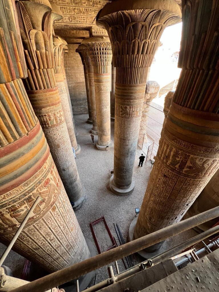 Interior view of the temple showing massive stone columns with detailed carvings and colorful hieroglyphs. The columns are shaped like lotus or papyrus plants and are closely spaced, supporting a decorated ceiling. Two people stand near the base of the columns, providing scale. The ground is covered in gravel, and scaffolding or railings are visible in the foreground.
