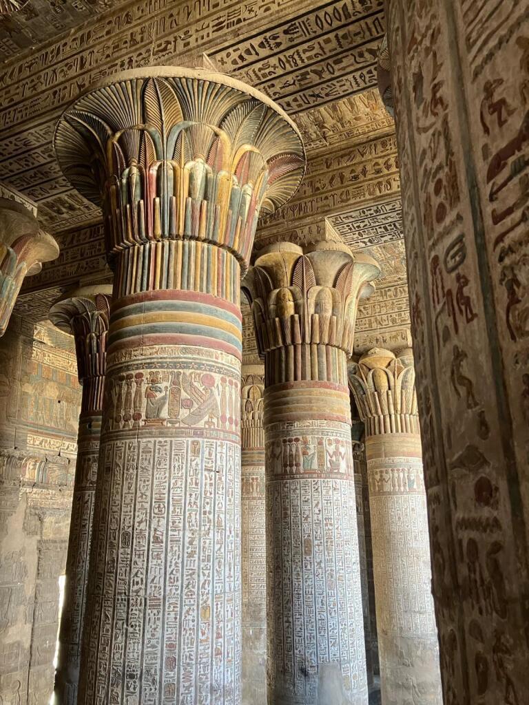 Interior view of rhe temple showing the ceiling and massive stone columns with detailed carvings and colorful hieroglyphs. The columns are shaped like lotus or papyrus plants and are closely spaced, supporting a decorated ceiling. 
