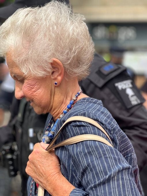An elderly lady with cottonwool white hair.  She's carrying a pale-coloured handbag, wearing a blue striped dress.  She's being arrested.