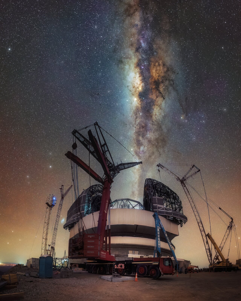 The image shows the open metallic dome of the Extremely Large Telescope (ELT) at the centre of the picture taking up the lower half of the frame. At the top, like a river the Milky Way band horizontally pours into the ELT. The construction site of the ELT is surrounded by cranes, while the Milky Way lights up the sky in various colours. One patch of the Milky Way shines the brightest, the centre of the galaxy.