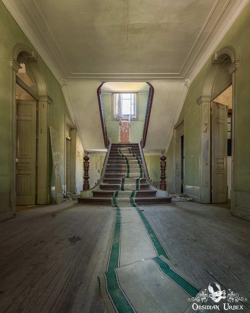 An abandoned grand foyer features a central staircase with peeling green runner, faded green walls, and natural light from a window above the stairs.