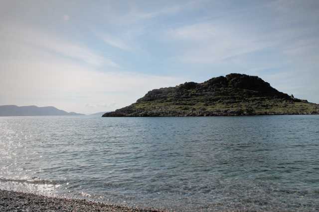 Das Bild zeigt eine ruhige Bucht mit kristallklarem Wasser und einem felsigen Hügel im Hintergrund. Der Himmel ist hellblau mit nur wenigen Wolken, was auf einen ruhigen und sonnigen Tag hindeutet. Das Wasser ist ruhig und reflektiert das Licht, während kleine Wellen sanft an den Kiesstrand schlagen. Die felsige Insel wirkt rau und unberührt, ein Kontrast zur ruhigen Atmosphäre des Wassers. Die Szene vermittelt ein Gefühl von Frieden, Ruhe und Einsamkeit, ein idealer Ort zur Entspannung und zum Genießen der Natur. Das Bild könnte ein Urlaubsfoto sein, das die Schönheit einer unberührten Landschaft einfängt. Die Farbpalette ist natürlich und beruhigend, mit sanften Blautönen im Wasser und den Grautönen des Felsens. Die Komposition ist einfach, aber effektiv, mit dem Fokus auf der Insel und dem Kontrast zwischen Land und Meer.